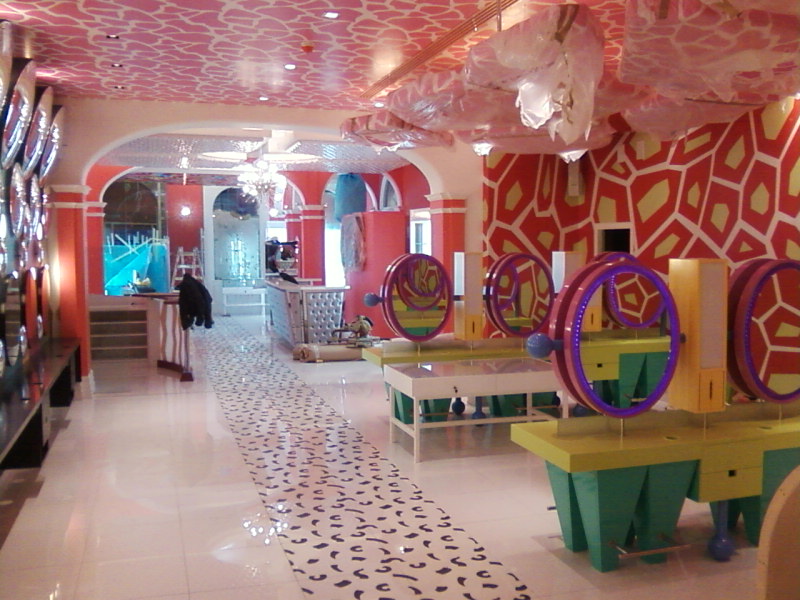 Wnętrze lokalu komercyjnego w trakcie wykańczania: ekscentryczny wystrój z wzorzystym sufitem, ścianami w geometryczne wzory i nietypowymi stanowiskami z okrągłymi lustrami, podłoga z wzorem...